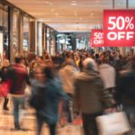 A bustling shopping center showcases a throng of shoppers eager for discounts. Bright sale signs promote savings as participants navigate through aisles carrying bags filled with purchases.