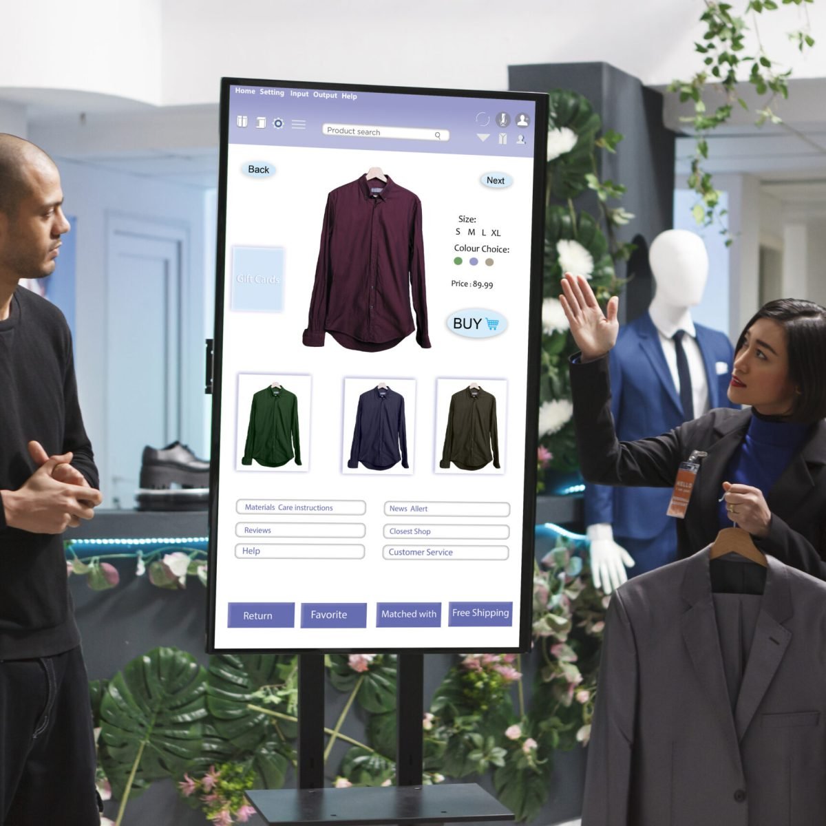 Asian woman holding a suit while assisting young man at the digital kiosk board with clothing prices. Female retail employee showing different clothes and merchandise to caucasian male client.